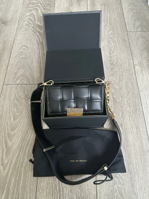 Svart quiltad handväska Ideal of Sweden - Snygg svart quiltad handväska från Ideal of Sweden med guldfärgade detaljer och bred axelrem. Väskan har en stilren rektangulär form och en dekorativ guldkedja. Tillverkad i konstläder och levereras med original dustbag och box. Nyskick. 