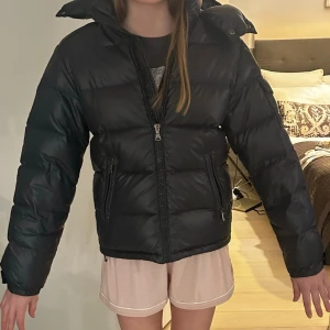 Svart dunjacka från Moncler - Snygg svart dunjacka från Moncler med hög krage och huva. Jackan har två dragkedjefickor fram och en Moncler-logga på ärmen. Puffig modell som håller värmen och ger en cool look. Perfekt för kalla vinterdagar. Helt äkta med QR kod, tyvärr har tvättlappen med animeringen inuti tyvärr åkt av dock.