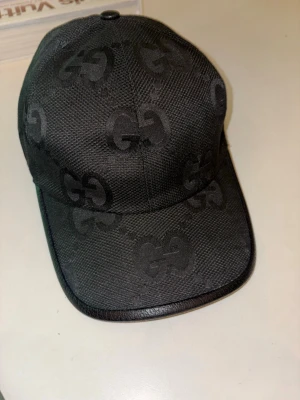 Svart keps med GG-mönster - Snygg svart keps med det klassiska GG-monogrammet över hela kepsen. Justerbar rem bak och detaljer i svart läder runt skärmen. Tillverkad i slitstarkt tyg som ger en exklusiv känsla. Perfekt accessoar för dig som gillar lyxiga streetwear-vibbar.