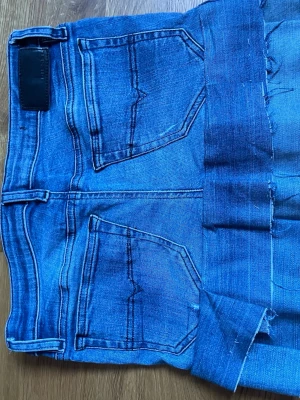 Blå jeanskjol med fransig kant - Snygg kort jeanskjol i blå denim med rå, fransig nederkant och klassiska fickor både fram och bak. Kjolen har knapp och dragkedja framtill samt tydliga sömmar för en cool vintagekänsla. Perfekt för dig som gillar avslappnad och trendig stil.