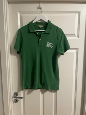Grön pikétröja från Burberry - Snygg grön pikétröja från Burberry med klassisk krage och knappar framtill. Tröjan har Burberrys ikoniska logga broderad på bröstet och är i slim fit-modell. Perfekt till jeans eller shorts för en clean och stilren look.