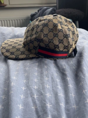 Gucci beige keps  - Snygg beige keps från Gucci med klassiskt GG-monogram över hela kepsen och en ikonisk röd och marinblå rand på sidan. Kepsen har böjd skärm och justerbar rem bak. Tillverkad i canvasmaterial som ger en exklusiv känsla och streetwear-vibe.
