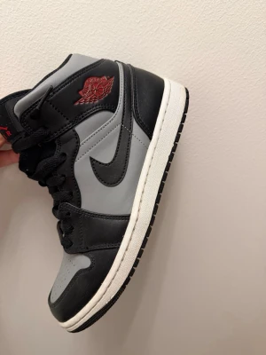 Air Jordan 1 Mid svart/grå - Snygga Air Jordan 1 Mid sneakers i svart och grått läder med klassisk Swoosh på sidan och röd Air Jordan-logga vid ankeln. Skon har svart snörning, vit mellansula och svart yttersula. Perfekt för dig som gillar streetwear och ikoniska modeller.