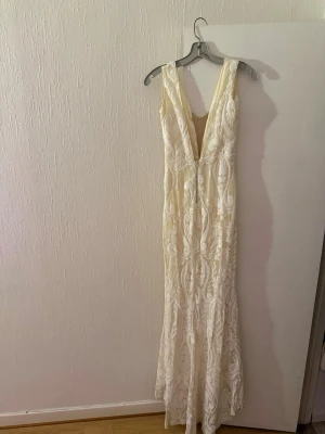 Vit långklänning med spets och paljetter - Elegant vit långklänning med djup v-ringning och vackra spetsdetaljer samt paljetter som ger extra lyster. Klänningen har figurnära passform och dragkedja i ryggen. Perfekt för speciella tillfällen när du vill sticka ut.