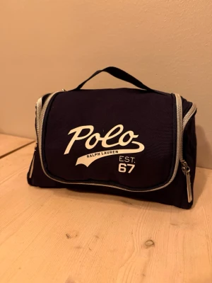 Marinblå necessär Polo Ralph Lauren - Snygg marinblå necessär från Polo Ralph Lauren med vit logga och text 'EST. 67' på framsidan. Väskan har dragkedja runtom, bärhandtag upptill och en praktisk krok för upphängning. Tillverkad i slitstarkt tygmaterial.