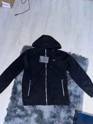 Svart vindjacka från Prada med huva - Snygg svart vindjacka från Prada med huva och dragkedja framtill. Jackan har två fickor med dragkedja och diskret Prada-logga på bröstet. Tillverkad i ett glansigt, lätt material som står emot vind. Perfekt för dig som vill ha en stilren och exklusiv look.
