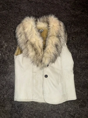 Vit pälsväst från BitchBabe - Säljer en snygg vit pälsväst från BitchBabe med fluffig fuskpälskrage i beige och grå toner. Västen har en knapp framtill och är fodrad med mjuk pälsimitation på insidan. Perfekt för dig som vill ha en statement piece till din outfit. (Mindre i storlek)