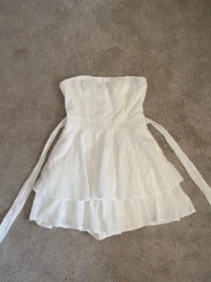 Vit axelbandslös volangklänning - Söt vit kortklänning med volanger och broderade detaljer. Klänningen är axelbandslös och har knytband i midjan för en snygg passform. Perfekt för sommarens alla tillfällen, exempelvis till studenten!🌷💝