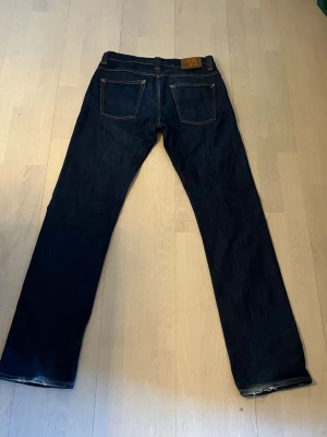 Mörkblå raka jeans från Nudie - Säljer ett par mörkblå jeans från Nudie (dry denim) med rak passform och klassiska fem fickor. Jeansen har kontrastsömmar och är tillverkade i kraftig denim. Perfekta för dig som gillar stilrena och tidlösa jeans.