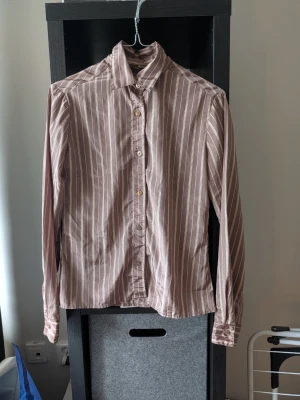 Brun och vit randig skjorta - Snygg brun skjorta med vita vertikala ränder och klassisk krage. Skjortan har långa ärmar och knäppning framtill med ljusa knappar. Perfekt för en stilren och avslappnad look.