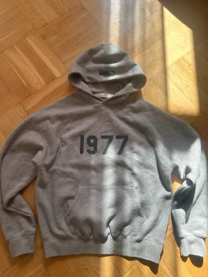 1977 Essentials Hoodie - Snygg grå hoodie med svart tryck '1977' på bröstet.  Använd 2 gånger men det märks inte. Sitter väldigt stor i storlek så XS sitter som S. Skriv om du har en fråga💫