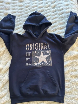 Mörkblå hoodie med stjärntryck - Mörkblå hoodie från Original Fit med stor vit stjärna och texten 'ORIGINAL FIT EST. 2025' framtill. Hoodien har en klassisk känguruficka och huva. Tillverkad i mjukt sweatshirtmaterial, perfekt för en avslappnad stil.