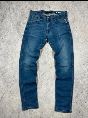 Replay anbass -  W32 L 32 Snygga blå jeans från Replay med klassisk femficksmodell och raka ben. Jeansen har en lätt tvättad look med subtila slitningar och kontrastsömmar. Märkespatch bak i midjan och detaljerade bakfickor. Perfekta för en avslappnad stil.