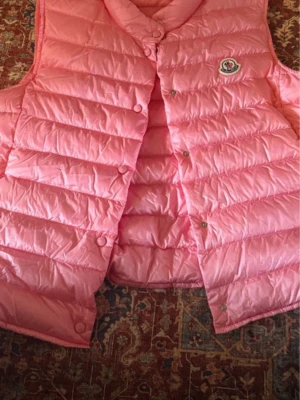 Rosa dunväst från Moncler - Säljer en snygg rosa dunväst från Moncler med quiltad design och tryckknappar framtill. Västen har en klassisk Moncler-logga på bröstet och är tillverkad i glansigt material som ger en fräsch look. Perfekt att slänga över en hoodie eller t-shirt för extra stil.