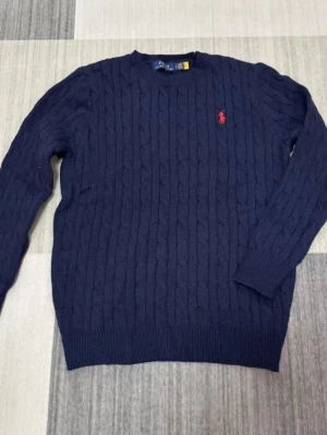 Mörkblå kabelstickad tröja Polo Ralph Lauren - Kabelstickad mörkblå tröja från Polo Ralph Lauren med rund halsringning och röd broderad logga på bröstet. Tröjan har långa ärmar och ribbade muddar vid ärmslut och nederkant. Perfekt för en klassisk och stilren look.