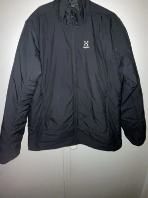 Svart vindjacka från Haglöfs - Snygg svart vindjacka från Haglöfs med dragkedja framtill och två sidofickor. Jackan har hög krage och diskret logga på bröstet samt vid nederkanten bak. Tillverkad i lätt och slitstarkt material, perfekt för blåsiga dagar.