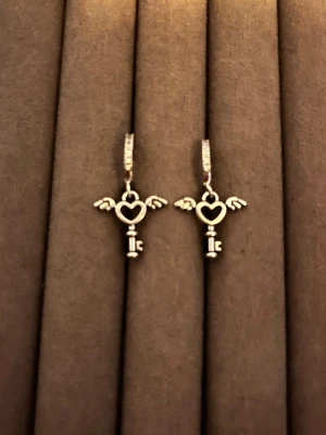 Silverörhängen med hjärtnyckel och vingar - Säljer ett par silverfärgade örhängen med hängande berlocker i form av nycklar med hjärtformad topp och små vingar på sidorna. Örhängena har gnistrande stenar på ringen och en unik, lekfull design som sticker ut. Perfekta för dig som gillar detaljer och coola accessoarer.