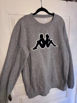 Grå sweatshirt från Kappa - Säljer en grå sweatshirt från Kappa med det klassiska logotrycket i svart och vitt på bröstet. Tröjan har rund halsringning och är tillverkad i mjukt sweatshirtmaterial, perfekt för en avslappnad och sportig stil.