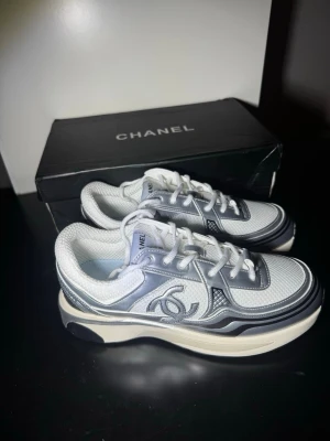 Vita och grå Chanel sneakers - Snygga sneakers från Chanel i vitt och grått med klassisk logga på sidan. Skorna har en chunky sula, mesh- och läderdetaljer samt vita skosnören. Perfekta för dig som vill ha en trendig och exklusiv look.