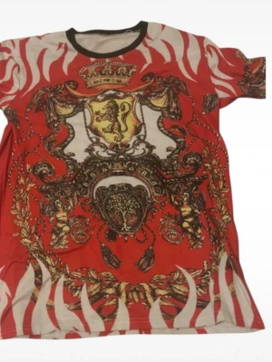 Röd t-shirt med lejon och barockmönster - Unik röd t-shirt med korta ärmar och ett stort barockinspirerat tryck med lejon, kronor och gulddetaljer. T-shirten har vita zebraliknande ränder och svart krage. Materialet känns som bomull eller polyester, perfekt för dig som gillar statement-plagg.