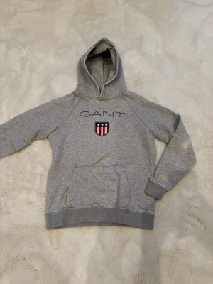 Grå hoodie från GANT med logga - Snygg grå hoodie från GANT med broderad logga och sköld på bröstet. Klassisk känguruficka framtill och mjukt material som känns skönt mot huden. Perfekt för dig som gillar stilrena och sportiga plagg. Dragsko i huvan saknas.