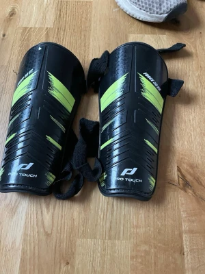 Pro Touch benskydd svart/grön - Säljer ett par benskydd från Pro Touch i svart med gröna detaljer. De har justerbara kardborreband för bra passform och en hård yta för skydd. Perfekta för fotbollsträning eller match.