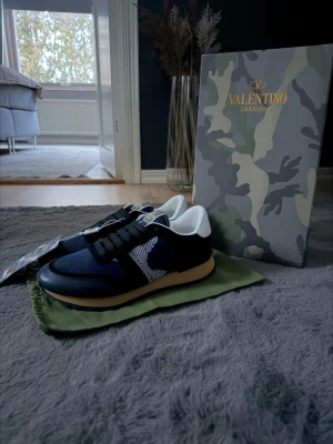 Valentino Garavani blå camo sneakers - Snygga sneakers från Valentino Garavani i blått och svart camo-mönster. Ovandelen är i mesh och mocka med vita detaljer och svart snörning. Skorna har en beige sula och vit insida. Perfekta för dig som vill sticka ut med exklusiv streetstyle.