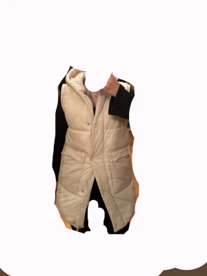 Beige lång dunväst från Zara - Snygg beige lång dunväst från Zara med huva och stora fickor framtill. Västen har quiltad design, tryckknappar och dragkedja. Perfekt för lager på lager och ger en trendig look till höst och vinter.