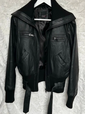 Som ny! Svart äkta läder jacka från CKN - Snygg svart äkta läder jacka från CKN med bred krage, ribbade muddar och midja samt bälte med spänne. Jackan har två bröstfickor med dragkedja och två snedställda fickor framtill. Perfekt för dig som vill ha en edgy och stilren look. Inköpt för ca 4000kr!