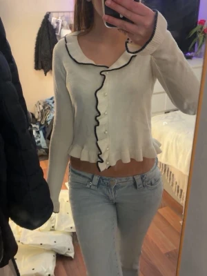 Vit kofta med volangkant och svarta detaljer - Söt vit kofta med volangkant och svarta vågiga detaljer längs knäppningen och halsringningen. Koftan har pärlknappar framtill och långa ärmar. Perfekt för en trendig och feminin look.