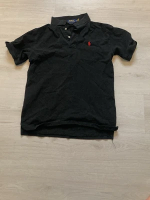 Svart pikétröja från Polo Ralph Lauren - Klassisk svart pikétröja från Polo Ralph Lauren med korta ärmar och krage. Tröjan har knappar vid halsen och den ikoniska röda logotypen broderad på bröstet. Perfekt för sommarn och varma dagar som kommer. (Priset är ej hugget i sten). 