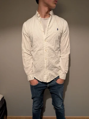 Vit skjorta från Ralph Lauren - Klassisk vit skjorta från Ralph Lauren med blå broderad logga på bröstet. Skjortan har lång ärm, knappar framtill och en tidlös passform. Perfekt till jeans eller chinos för en clean och stilren look.