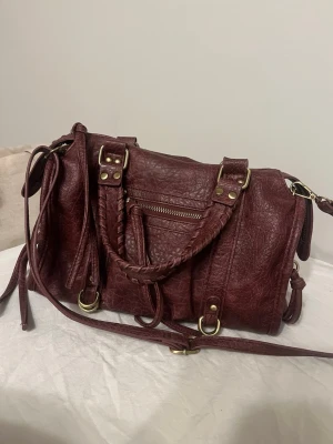 Populär Vinröd handväska från bershka  - Snygg vinröd handväska i läder med flätat handtag, justerbar axelrem och guldfärgade metalldetaljer. Perfekt för dig som vill ha en trendig och rymlig väska med coola detaljer. Populär väska från bershka. Inte använt mycket. 