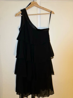 Svart oneshoulder volangklänning - Säljer en svart kortklänning med oneshoulder-design . Klänningen har flera lager volanger som ger ett snyggt och livfullt intryck. Perfekt för dig som vill sticka ut med en unik och trendig look.