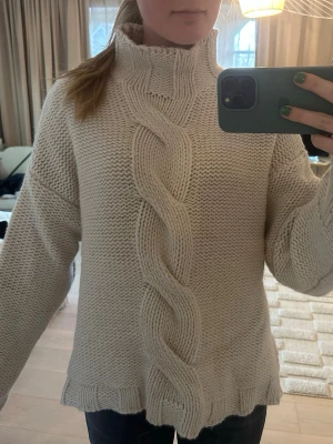 Beige kabelstickad polotröja  - Mysig beige stickad tröja med hög polokrage och en bred fläta framtill. Tröjan har slitsar i sidorna, ribbade muddar och är oversized i modellen. Perfekt för kalla dagar och enkel att styla med jeans.