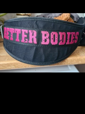 Svart träningsbälte Better Bodies - Säljer ett svart träningsbälte från Better Bodies med bred kardborrestängning och rosa broderad logga. Bältet är tillverkat i slitstarkt tyg och ger extra stöd vid tunga lyft på gymmet. Perfekt för dig som vill ha stabilitet och stil under träningen.