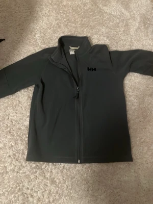 Mörkgrå fleecejacka Helly Hansen - Mjuk och varm mörkgrå fleecejacka från Helly Hansen med hel dragkedja framtill och hög krage. Diskret HH-logga på bröstet. Perfekt för lager-på-lager och outdoor-aktiviteter. Tillverkad i skön fleece som håller värmen.