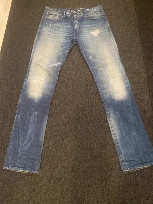 Blå slitna jeans från Replay - Säljer ett par blå jeans, We Are Replay, med slitningar och ljusa partier på knän och lår. Klassisk femficksmodell med raka ben och broderad bakficka. Jeansen har en cool, använd look och är tillverkade i bomull. Passar både kvinnor och män!