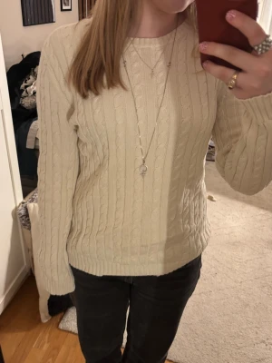 Vit kabelstickad tröja från Ralph Lauren - Säljer en klassisk vit kabelstickad tröja från Lauren Ralph Lauren. Tröjan har rund halsringning, långa ärmar och ribbade muddar vid ärmslut och nederkant. Perfekt för lager-på-lager och stilrena outfits.