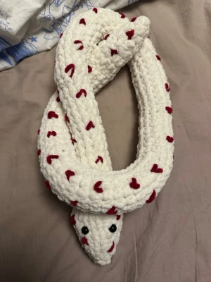 Vit stickad  orm hjärtan - Unik handgjord orm i mjukt, vitt garn dekorerad med små röda hjärtan längs hela kroppen. Ormen har svarta ögon och är perfekt som mysig prydnad eller kramdjur. Passar dig som gillar gulliga och personliga detaljer i rummet. Nästan 1 meter lång när den är uträttad 