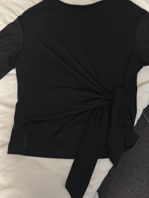 Svart topp med knyt i midjan - Snygg svart topp från H&M med lång ärm och trendig knytning i midjan. Perfekt för dig som gillar stilrena plagg med en twist. Tillverkad i mjukt material som känns skönt mot huden.