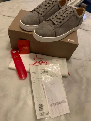 Gråa sneakers från Christian Louboutin - Snygga gråa sneakers från Christian Louboutin i mocka med klassisk röd sula och vita kanter. Lågt skaft och stilren design med matchande grå skosnören. Perfekta för dig som vill ha något exklusivt och trendigt i garderoben.