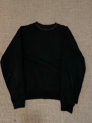 Svart sweatshirt från Acne Studios - Säljer en svart sweatshirt från Acne Studios i storlek XXS. Tröjan har rund halsringning med Acne Studios-logga runt kragen och är tillverkad i mjuk bomull. Perfekt för en clean och stilren look.