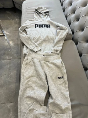 Grå tracksuit (hoodie) Puma med logga pojke/flicka - Ljusgrå tracksuit från Puma i mjukt sweatshirtmaterial. Stor svart Puma-logga på bröstet, klassisk huva och magficka. Ribbade muddar vid ärmslut och nederkant ger en clean och sportig vibe. Storlek 13-14
