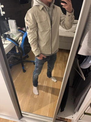 Beige jacka från C.P. Company - Säljer en beige jacka från C.P. Company med svart dragkedja och ikonisk linsdetalj på ärmen. Jackan har en ficka på ärmen. Perfekt för dig som gillar stilren och modern design.