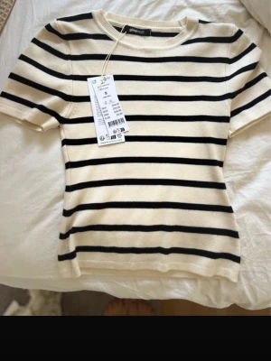 Randig topp från Gina Tricot - Säljer en krämvit och svart randig topp från Gina Tricot. Toppen har rund halsringning, korta ärmar och är tillverkad i mjuk bomullsmix. Perfekt för en enkel och stilren look.