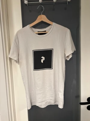 Vit t-shirt med svart tryck - Vit t-shirt från Peak Performance med ett svart fyrkantigt tryck och logga på bröstet. Klassisk rund halsringning och korta ärmar. Tillverkad i mjuk bomull för en skön känsla och enkel stil.