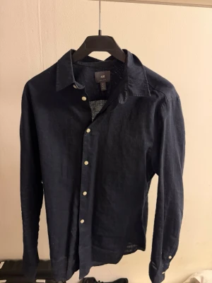 Mörkblå linneskjorta från H&M - Snygg mörkblå skjorta från H&M i luftigt linne. Klassisk krage, vita knappar framtill och långärmad modell. Perfekt för dig som gillar en stilren och enkel look med en relaxed vibe.