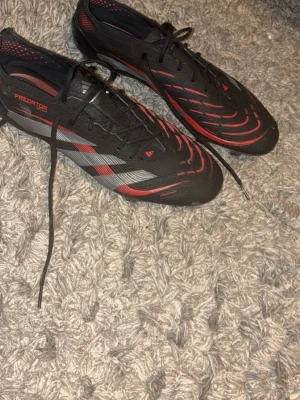 Adidas Predator svart/röd fotbollssko - Säljer ett par Adidas Predator fotbollsskor i svart med röda och grå detaljer. Skorna har snörning, låg profil och en modern design med mönstrad ovandel i syntetmaterial. Perfekta för dig som vill ha kontroll och stil på planen. Strl 46 2/3 kan gå ner i pris vid snabb affär, använda 1 gång!