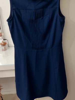 Marinblå ärmlös klänning - Snygg marinblå klänning i kort modell med ärmlös design. Framtill finns dekorativa vertikala veck som ger en stilren touch. Klänningen har en enkel, klassisk siluett och är tillverkad i ett mjukt tyg som känns bekvämt mot huden.